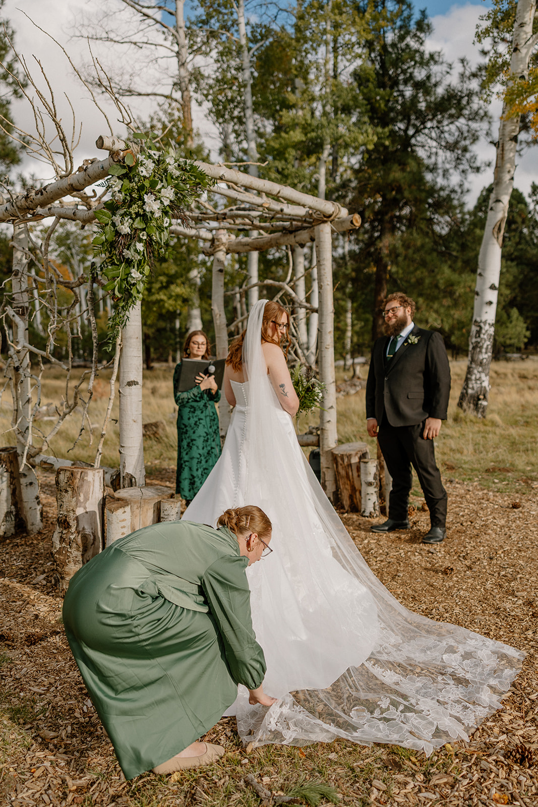 Bridesmaid adjusts the bride’s lace train as she stands at the altar with her groom beneath a birchwood arbor.