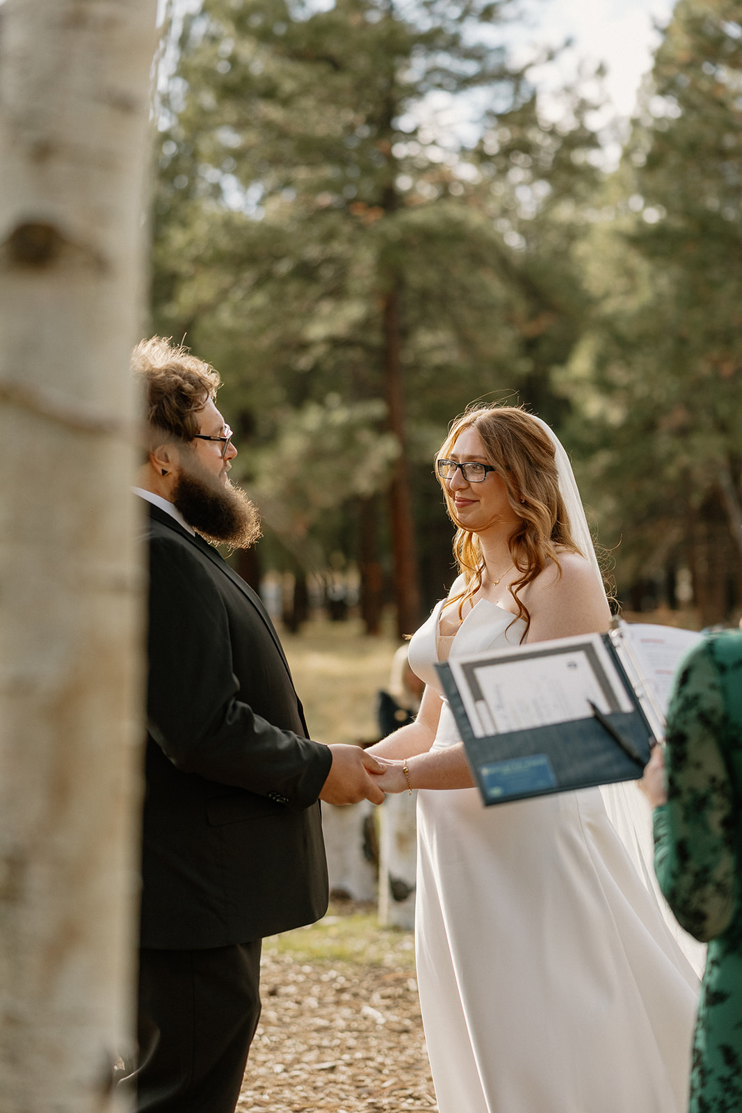 Bride and groom hold hands and exchange vows during their outdoor ceremony surrounded by tall pine trees.