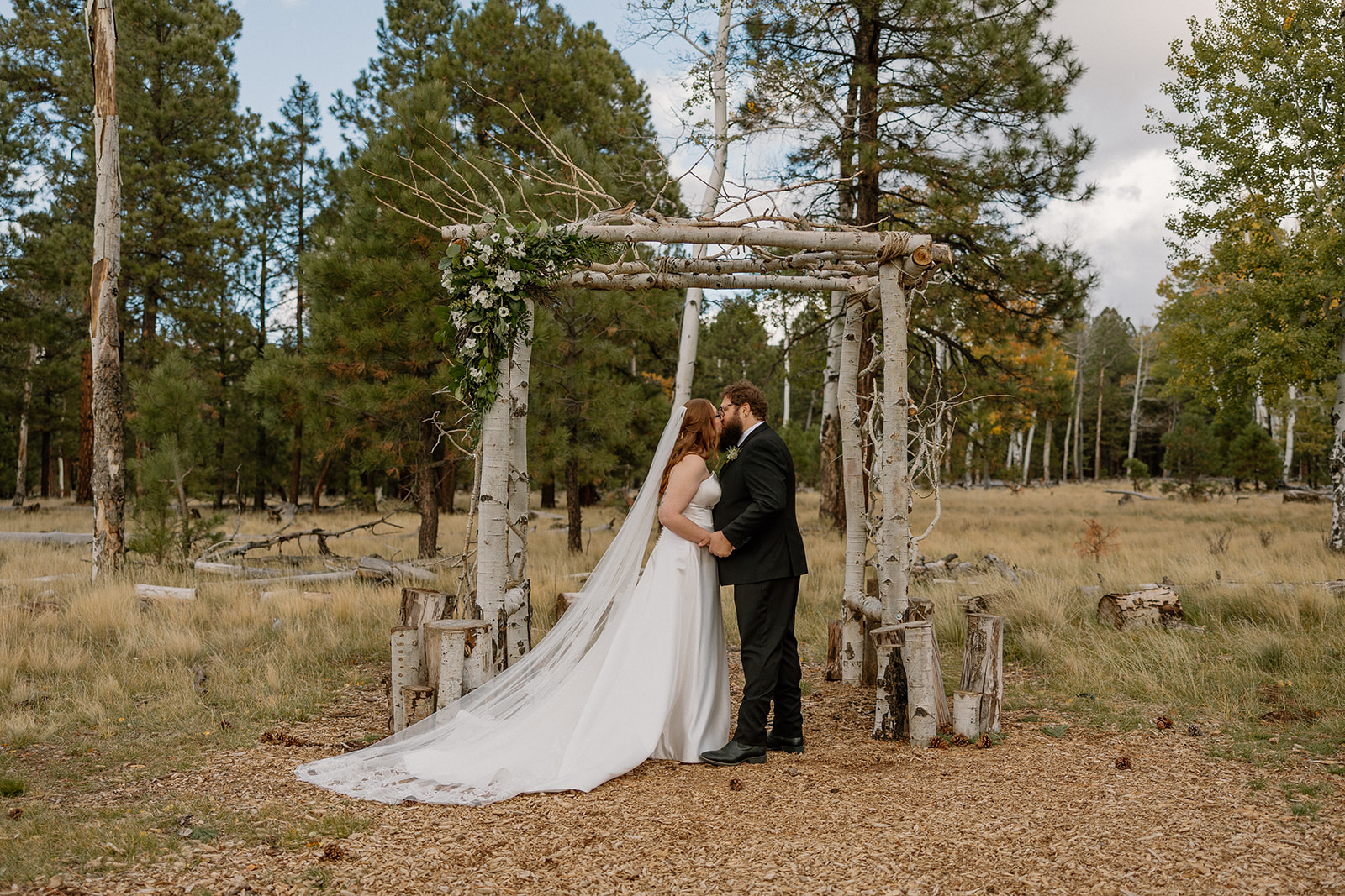 Bride and groom’s first kiss under a rustic birch arbor, surrounded by golden grass and autumn trees at Nordic Village AZ.