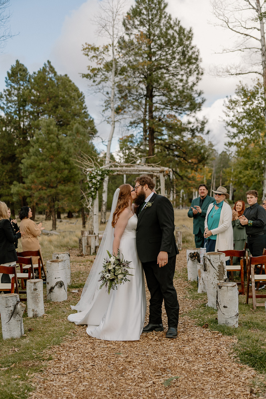 Newlyweds share a kiss at the end of the aisle, guests cheering behind them in the Nordic Village AZ woods.