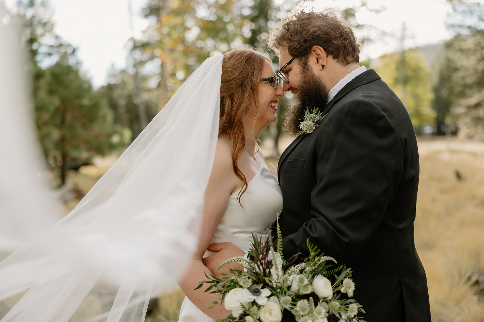 Bride and groom share a forehead-to-forehead moment in the woods, her veil floating in the breeze as they smile, surrounded by tall pines at Nordic Village AZ.