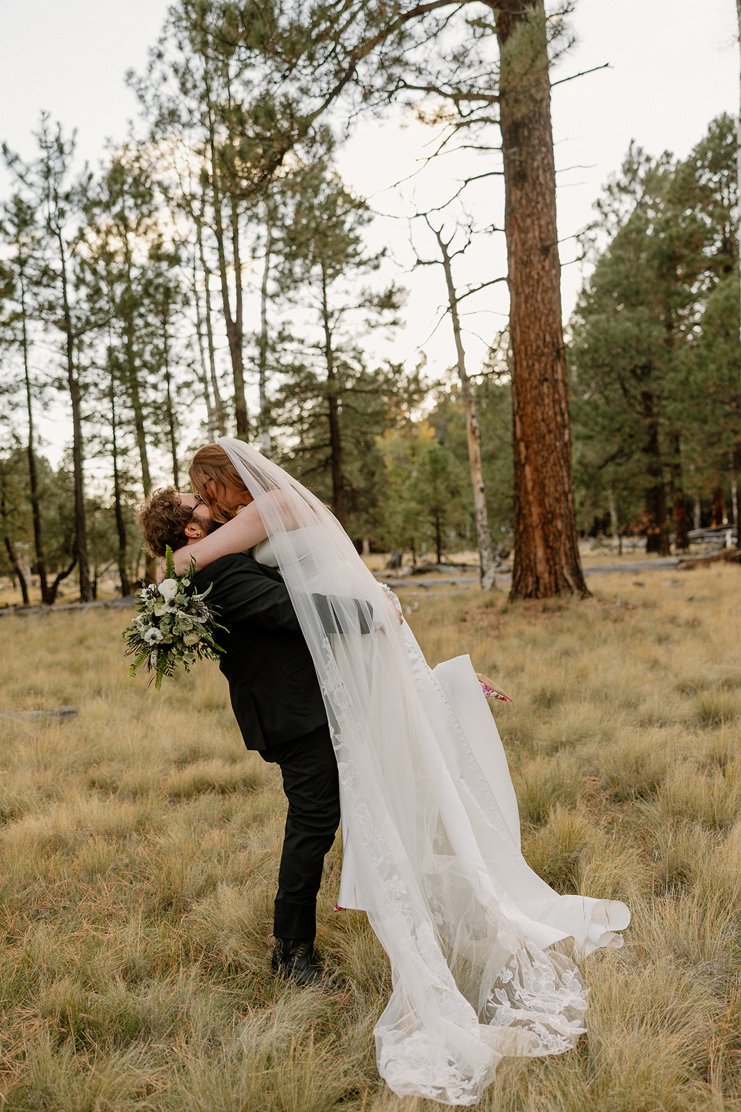 Bride balances on a fallen log while holding her groom’s hand, sunlight filtering through the trees at their Nordic Village AZ wedding.