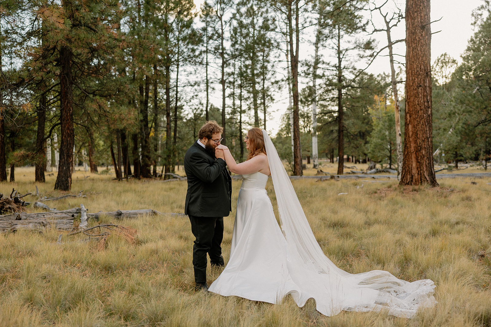 Groom kisses the bride’s hand in a soft, romantic moment among the pines and golden grass at Nordic Village AZ.