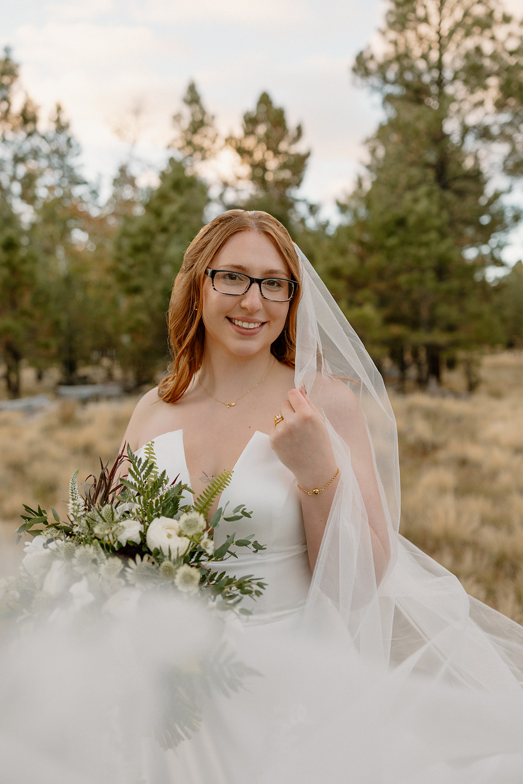 Bride smiles softly while holding her veil and bouquet, standing in a sunlit clearing with pine trees behind her.