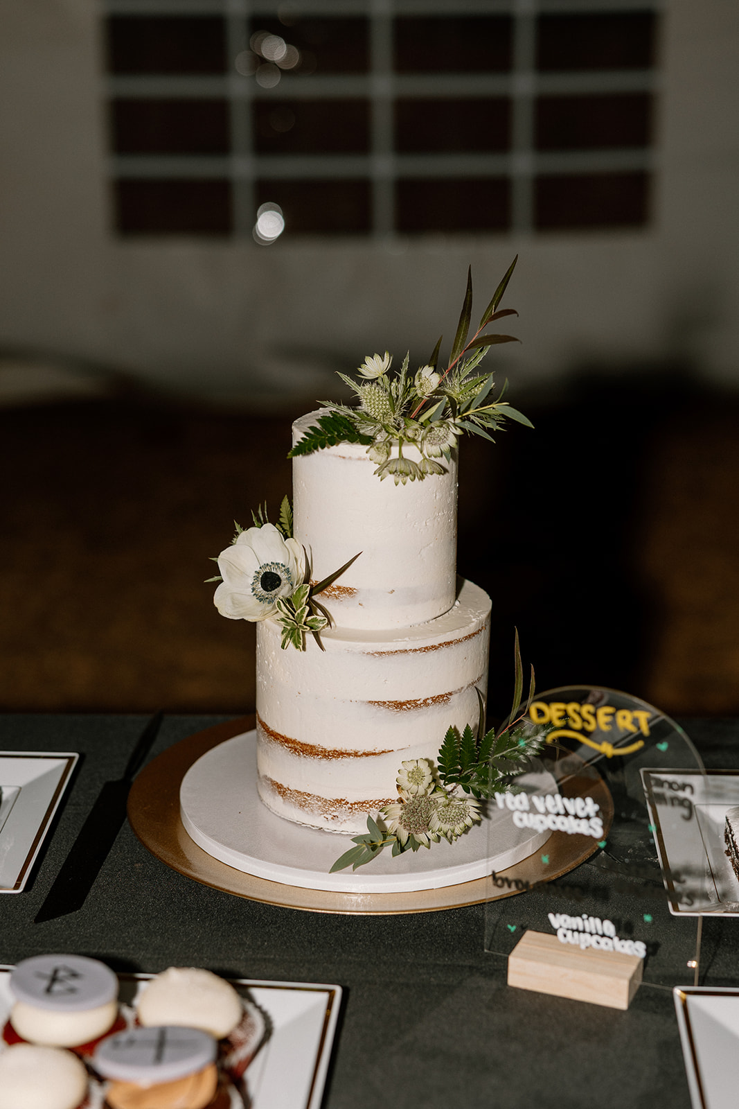Minimalist two-tier semi-naked wedding cake decorated with greenery and white blooms, styled with a dessert bar setup.