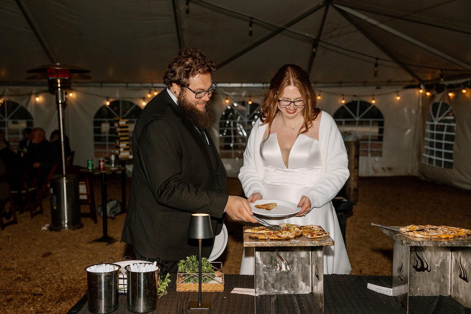 Bride and groom serve up slices of wood-fired pizza under the reception tent, laughing as they enjoy their late-night snack.