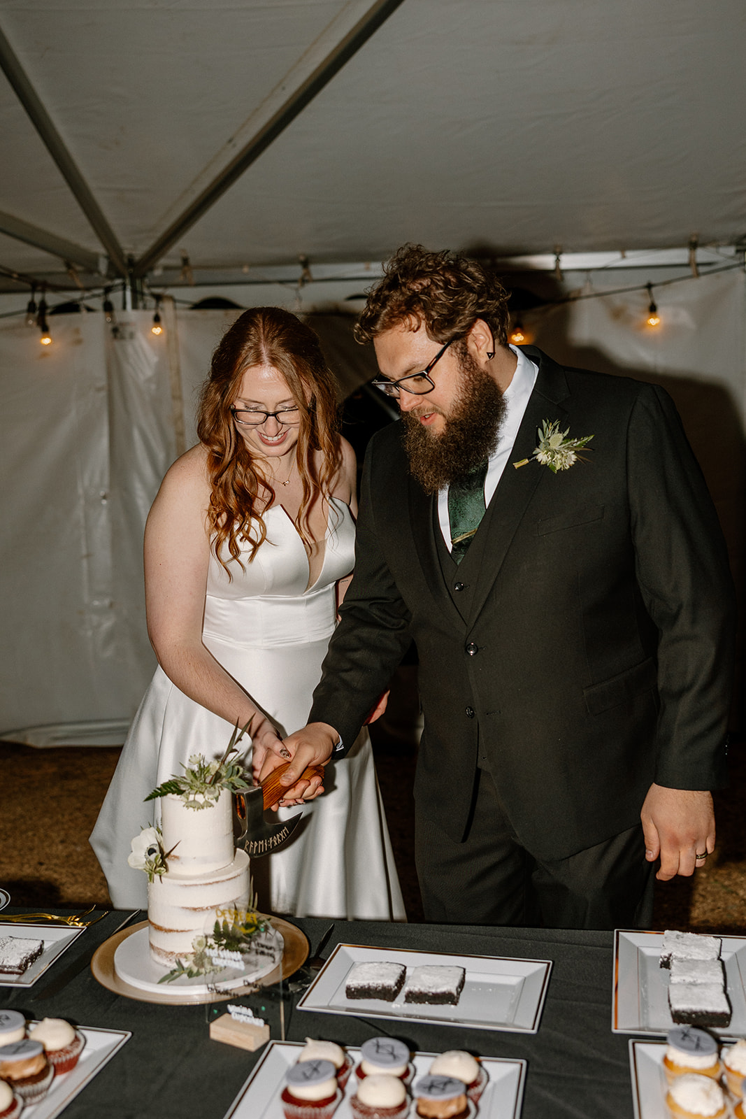Couple cuts their wedding cake together under a warmly lit tent, surrounded by trays of cupcakes and desserts.