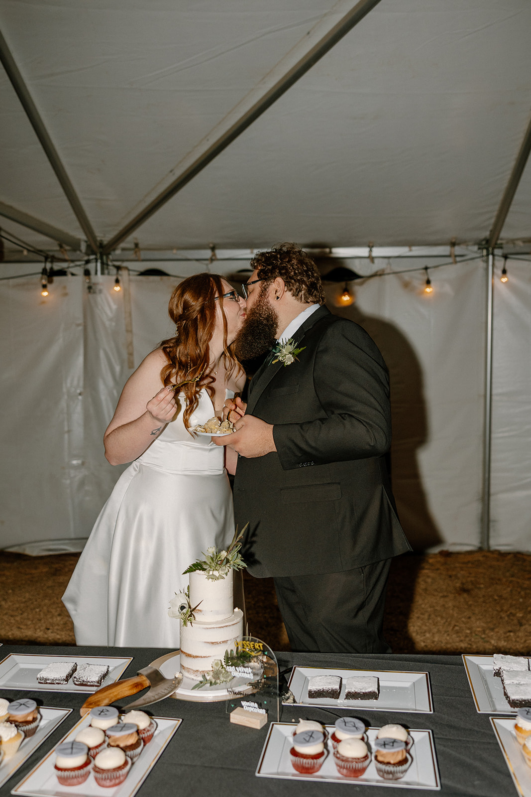 Bride and groom share a kiss behind their wedding cake after slicing into the first piece, joyful and sweet under the tent lights.