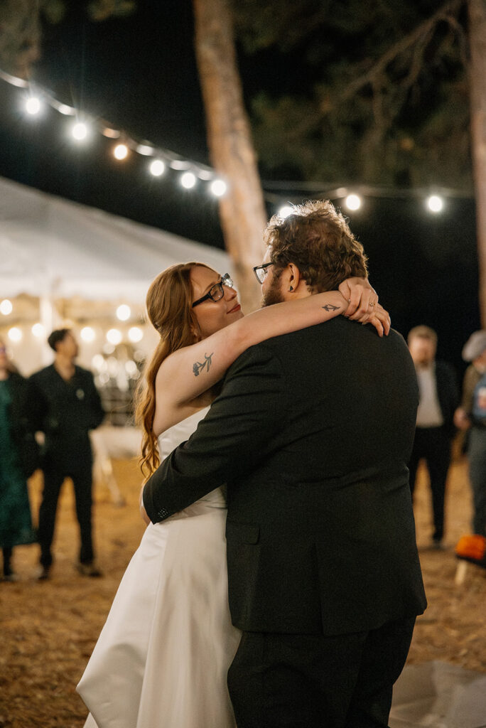 Groom embraces the bride during their first dance, her tattooed arms wrapped around him beneath strings of lights at Nordic Village AZ.