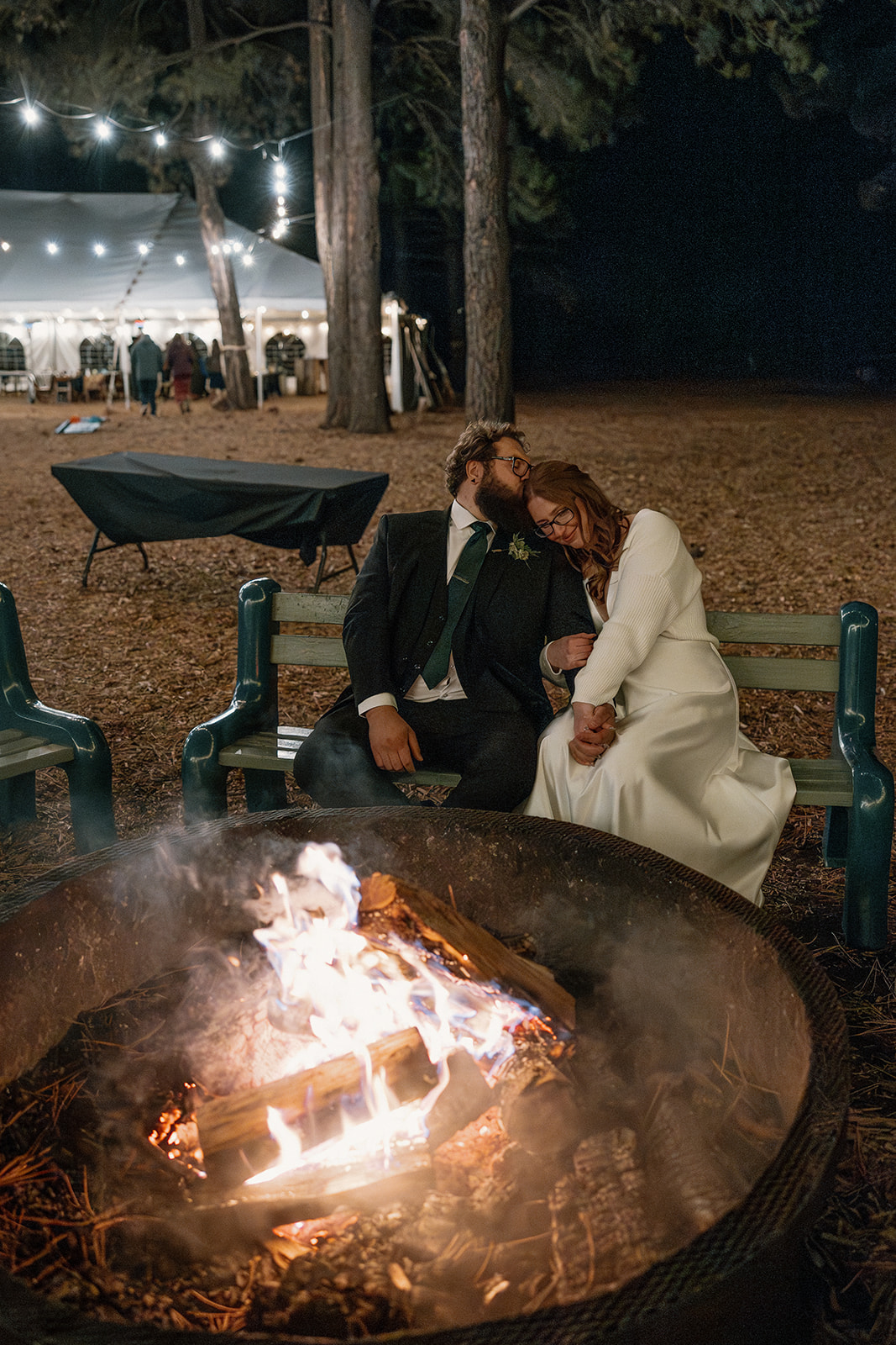 Couple cozies up by a glowing fire pit under string lights, with a white tent lit in the background at Nordic Village AZ.