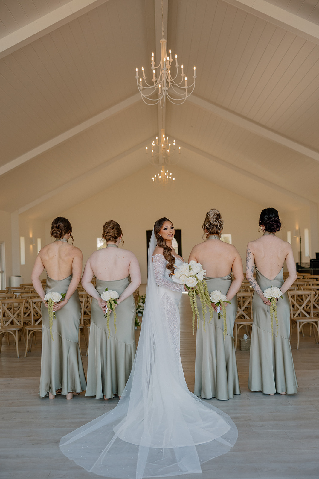 Back view of bride and bridesmaids inside a modern venue, showcasing satin dresses and soft florals.