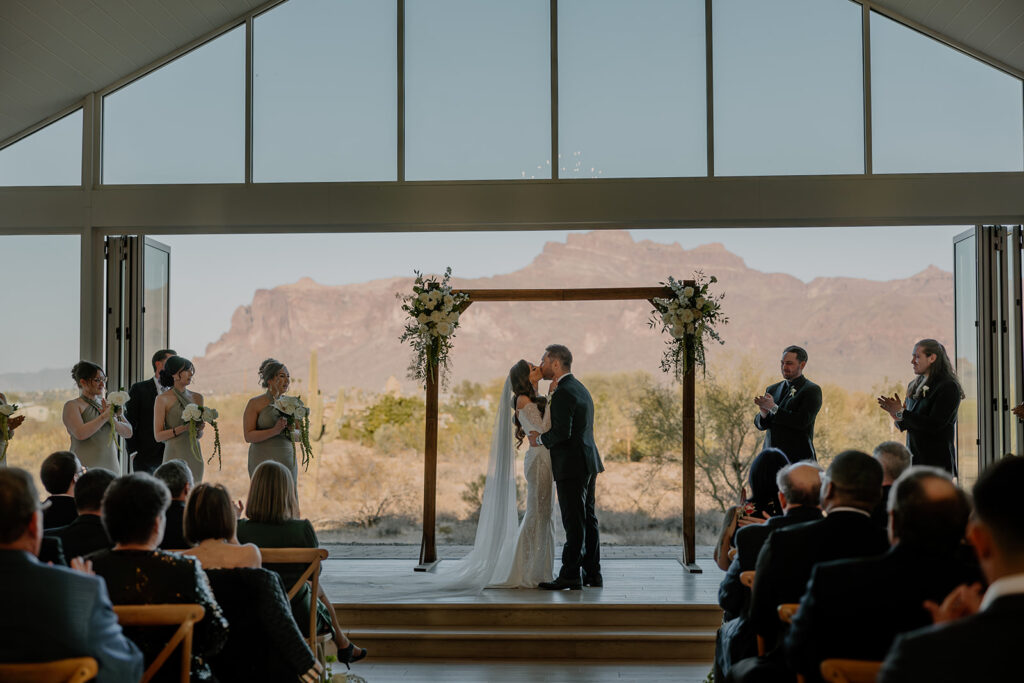 Bride and groom share their first kiss under a floral arch with Arizona mountains in the distance — perfect for your wedding planning guide vision board.