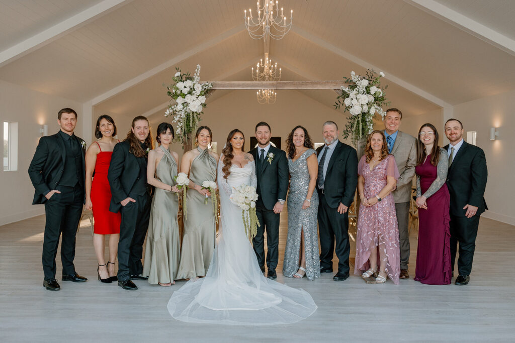 Extended family photo in a bright ceremony space with white florals and modern chandeliers.