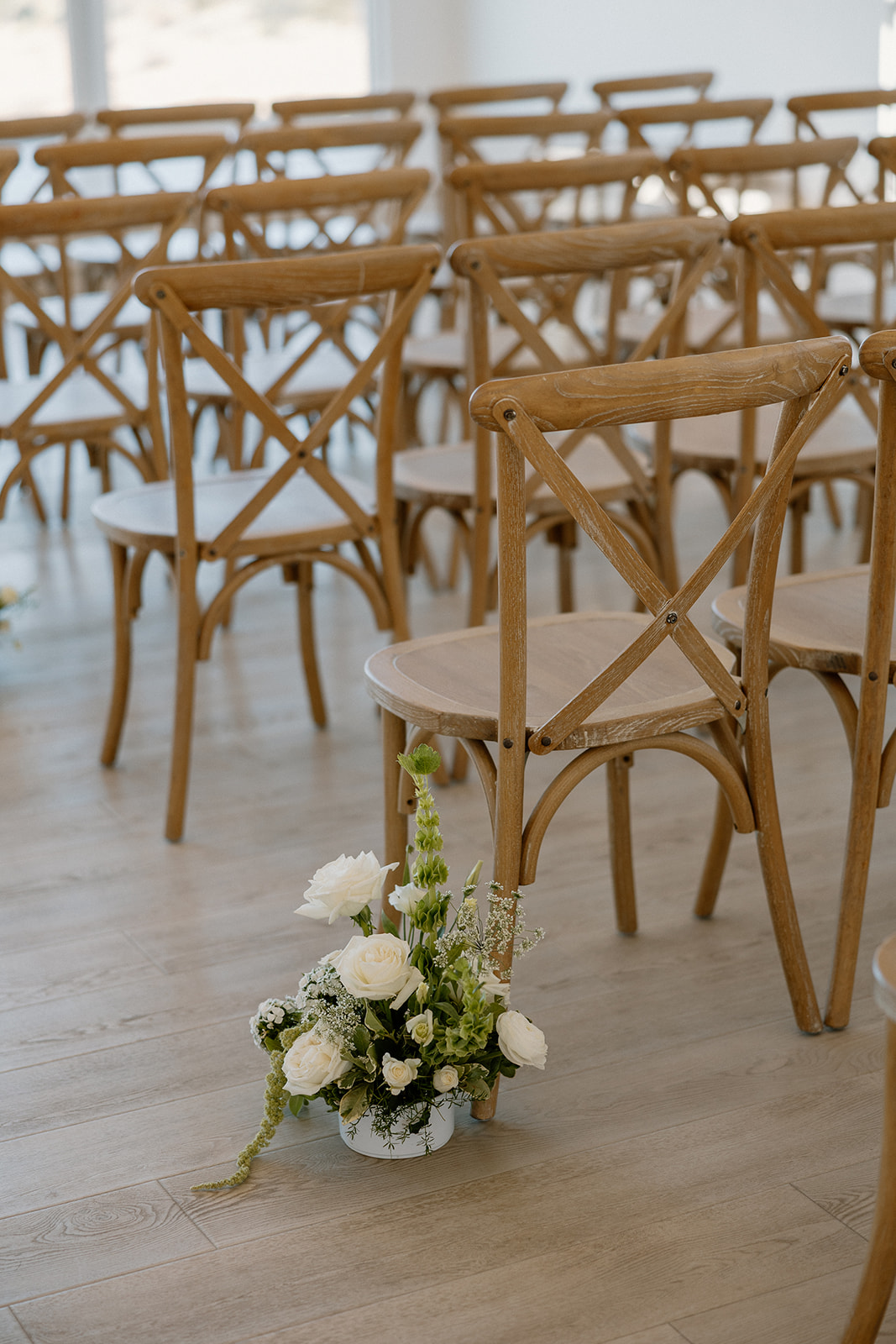 Natural wood chairs and white floral arrangements line the ceremony aisle in a minimalist indoor venue.