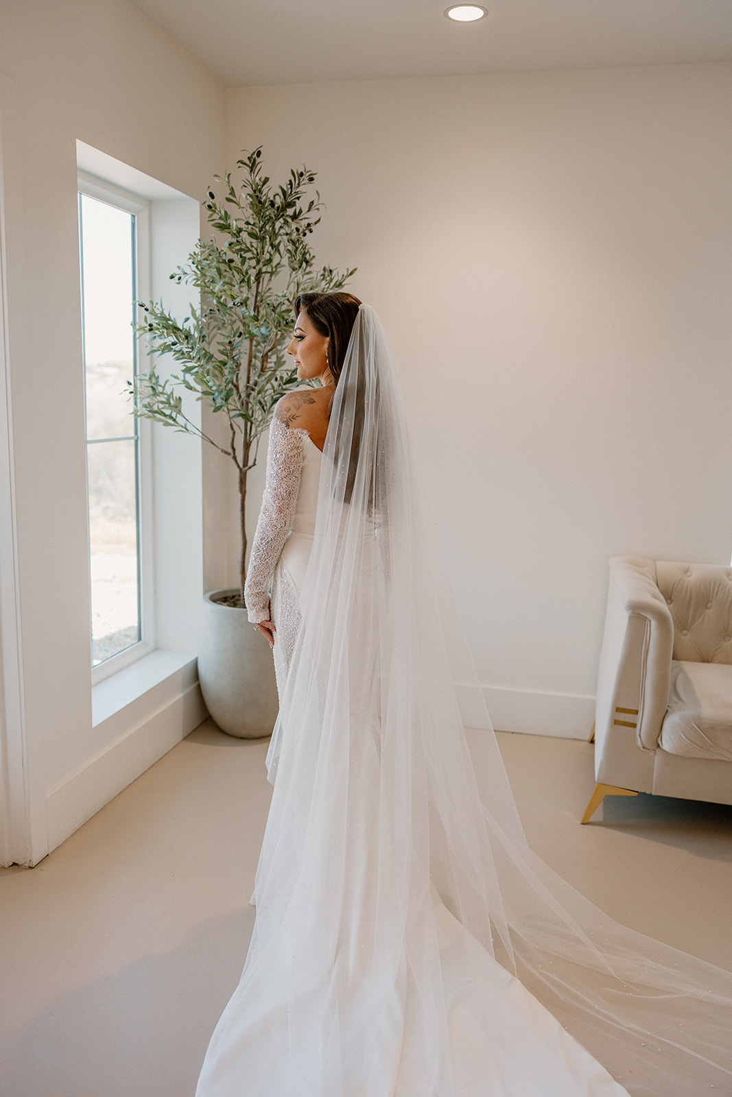 Back view of a bride in a fitted gown with a dramatic train, standing in a sunlit getting-ready space.