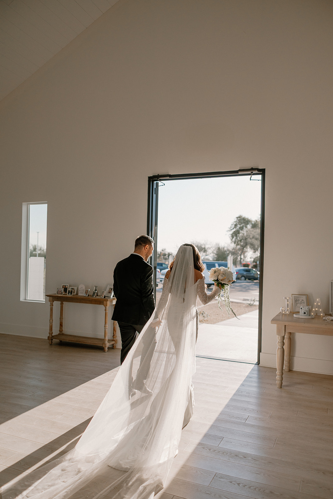 Bride and groom walking into a sunlit indoor space, her long veil trailing behind as they head toward the open doors.