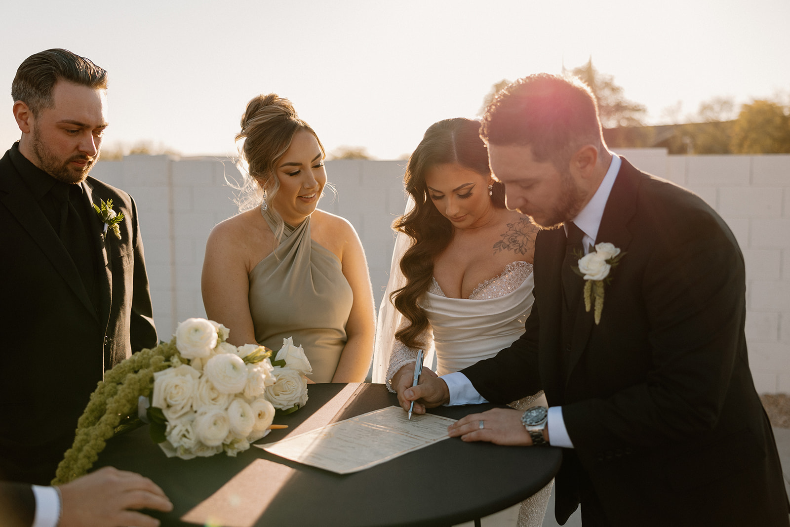 Bride and groom signing their marriage license outdoors, surrounded by members of their wedding party and soft floral arrangements.