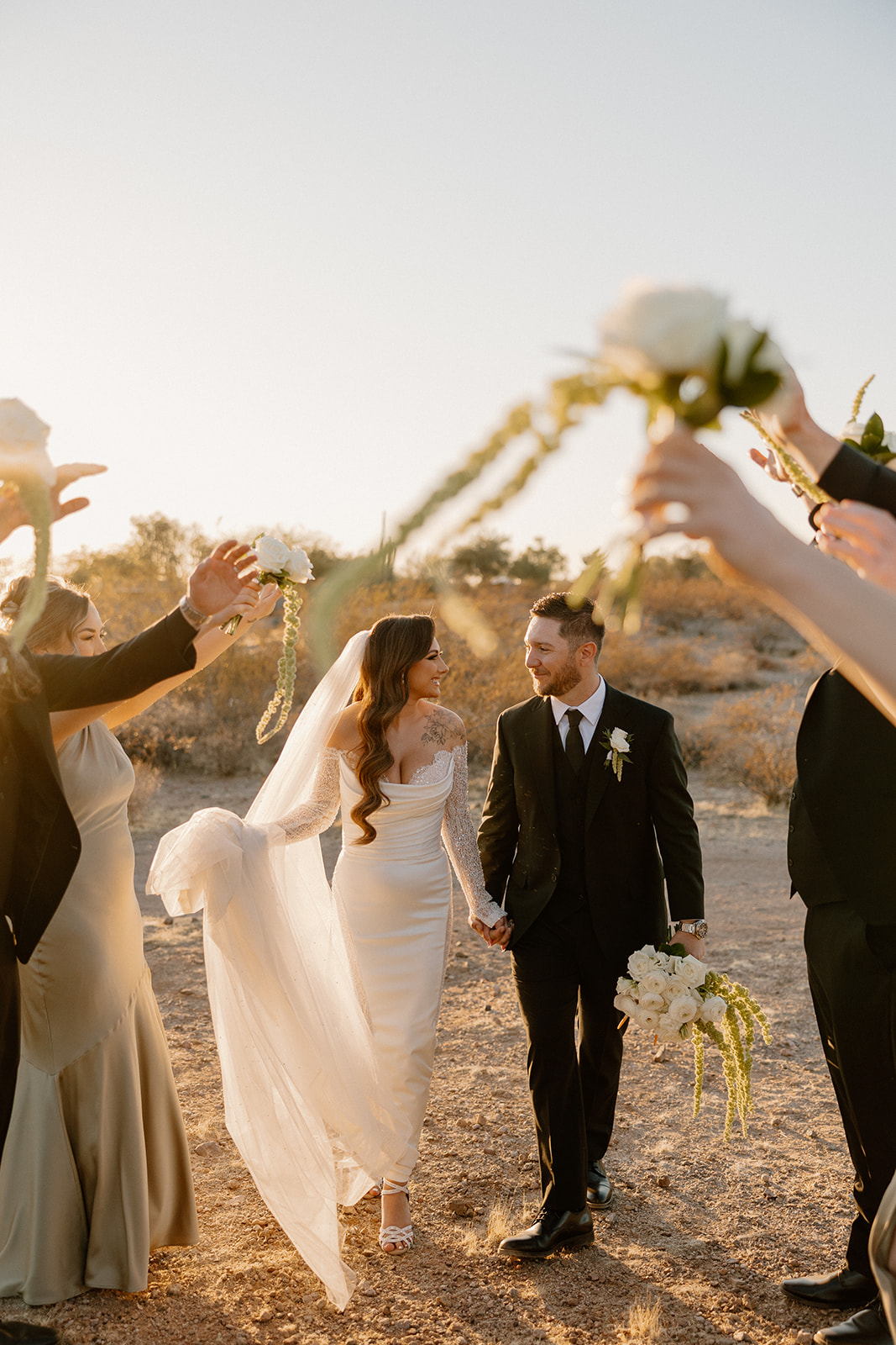 Bride and groom walk hand-in-hand through a celebratory tunnel of bridesmaids and groomsmen, golden light highlighting the desert setting.