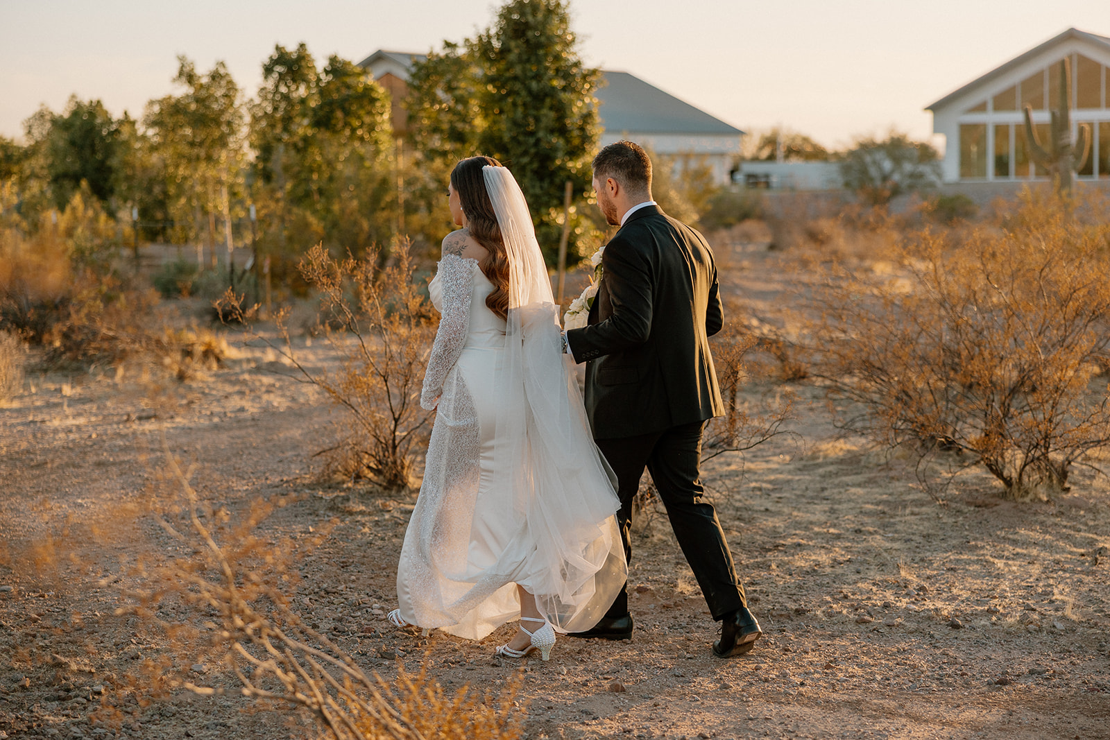 Bride and groom walk side-by-side through soft desert brush in the late afternoon glow, her veil trailing behind.