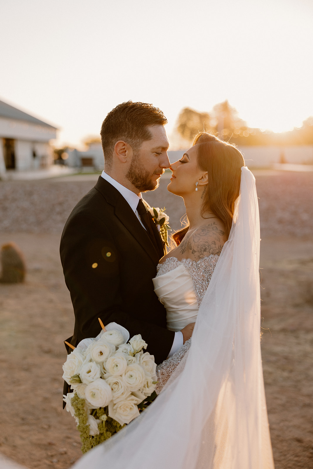 Bride and groom gaze at each other, arms wrapped close, standing on desert ground just before sunset at a desert view wedding venue.