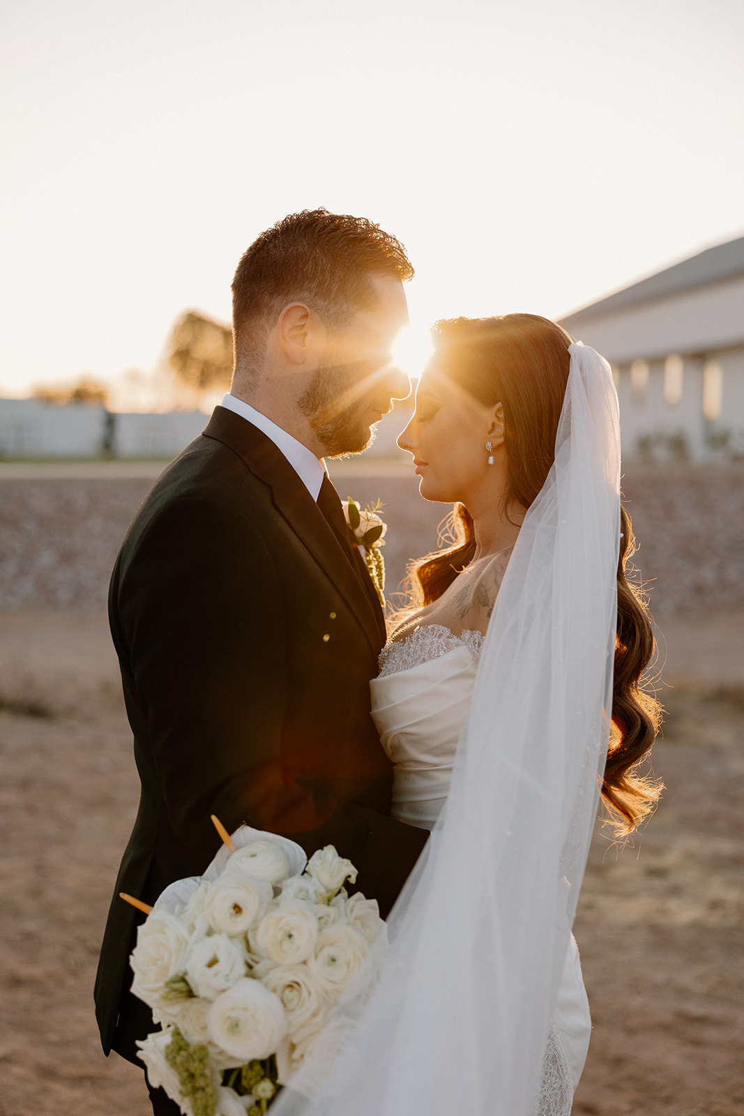 Bride and groom stand forehead to forehead, the sun peeking between them, wrapped in golden light and desert charm outside their wedding venue.