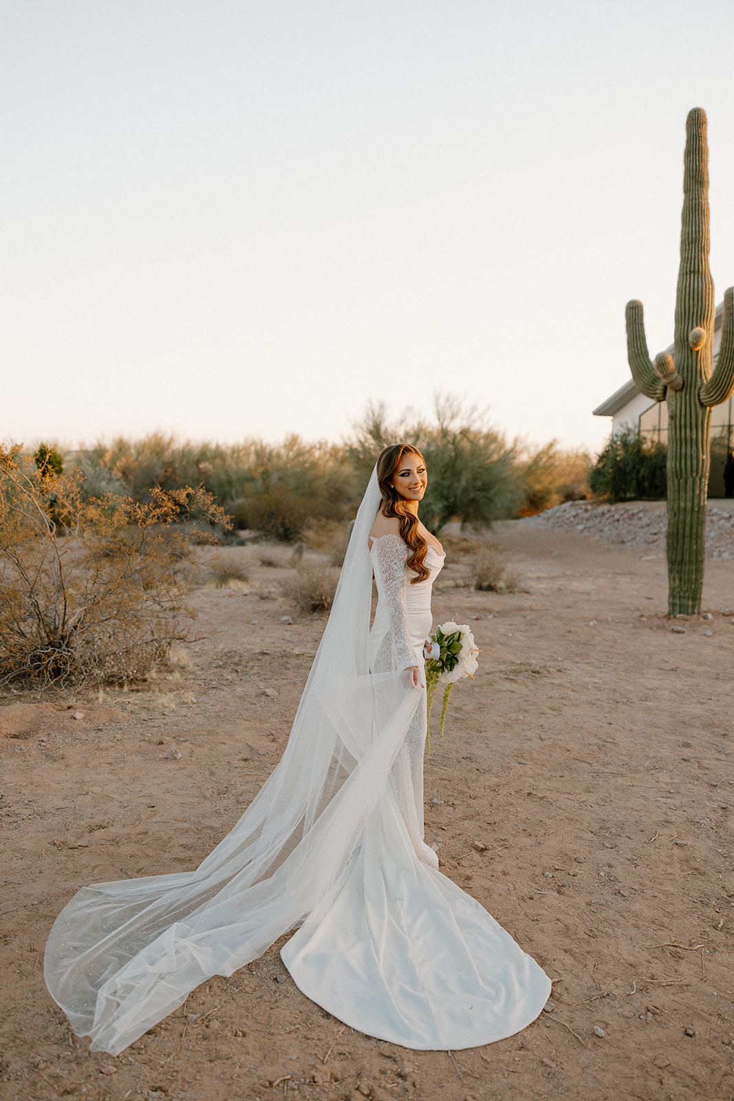 Bride stands with her long veil trailing behind her, bouquet in hand, against the desert backdrop of a cactus and her wedding venue.