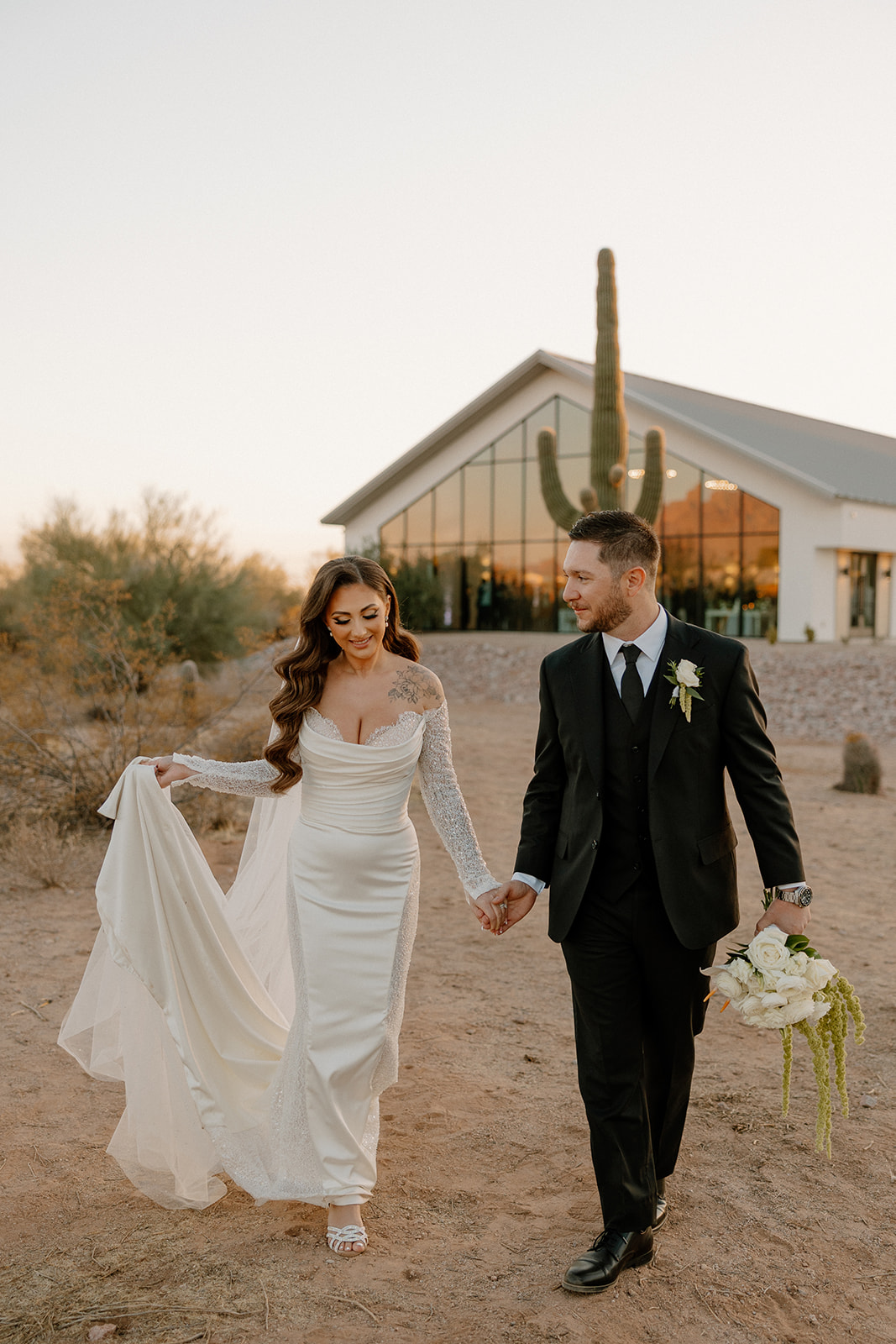 Bride lifts her dress as she walks beside her groom, a tall cactus and the modern venue structure behind them at a desert view wedding venue.