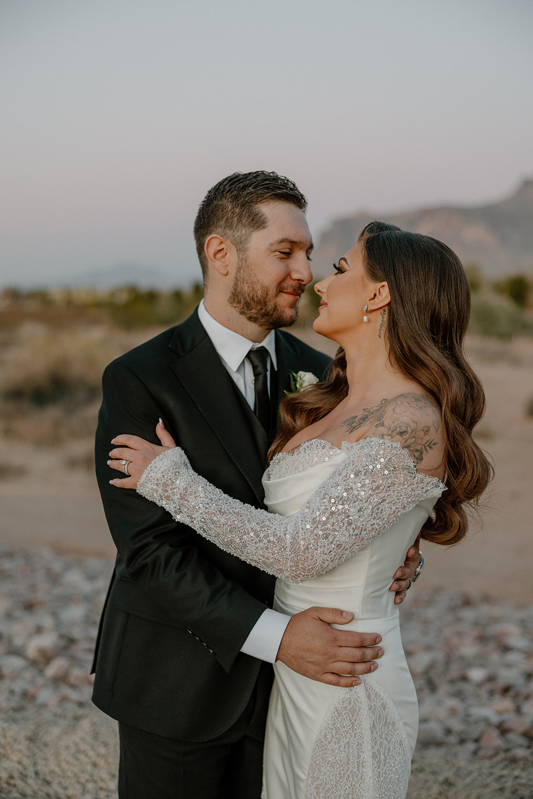 Groom holds the bride close as she smiles up at him, sunlight catching her veil and white rose bouquet outside their Arizona venue.