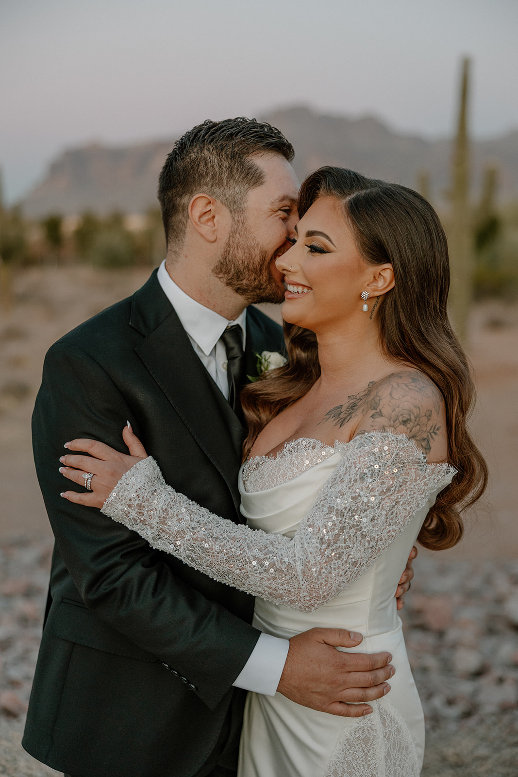 Bride and groom embrace and smile cheek-to-cheek, the desert landscape and cacti softly blurred in the background at a desert view wedding venue.