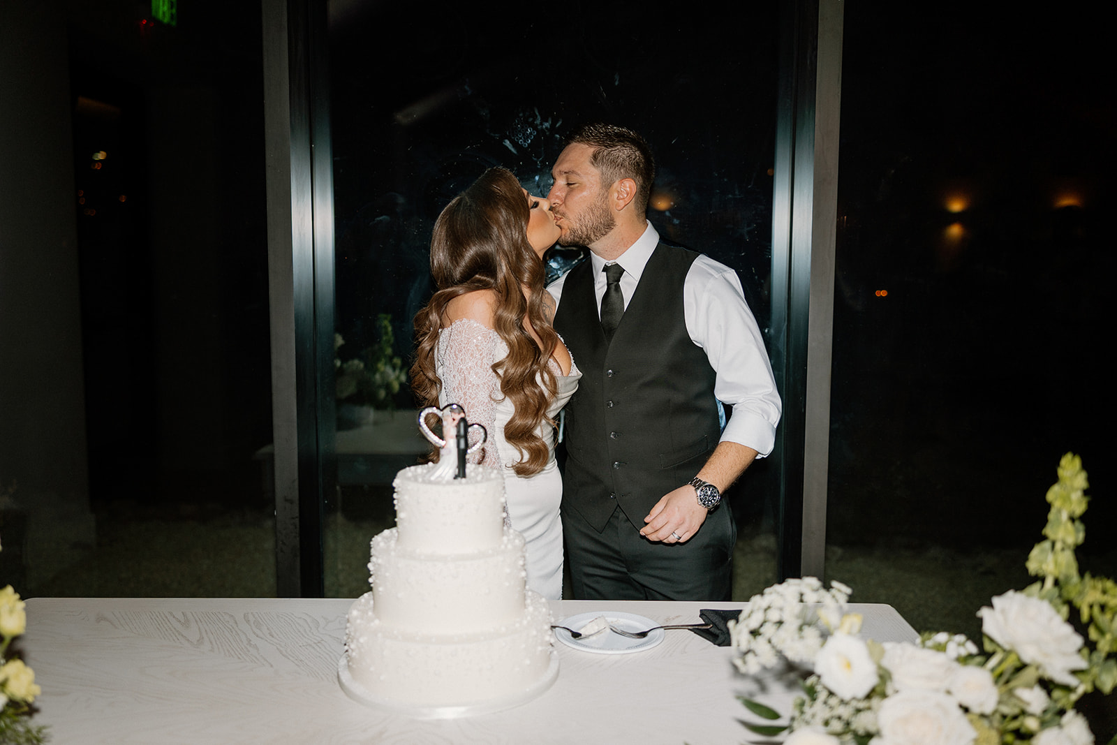 Bride and groom share a kiss in front of their three-tier white wedding cake during their evening reception indoors.