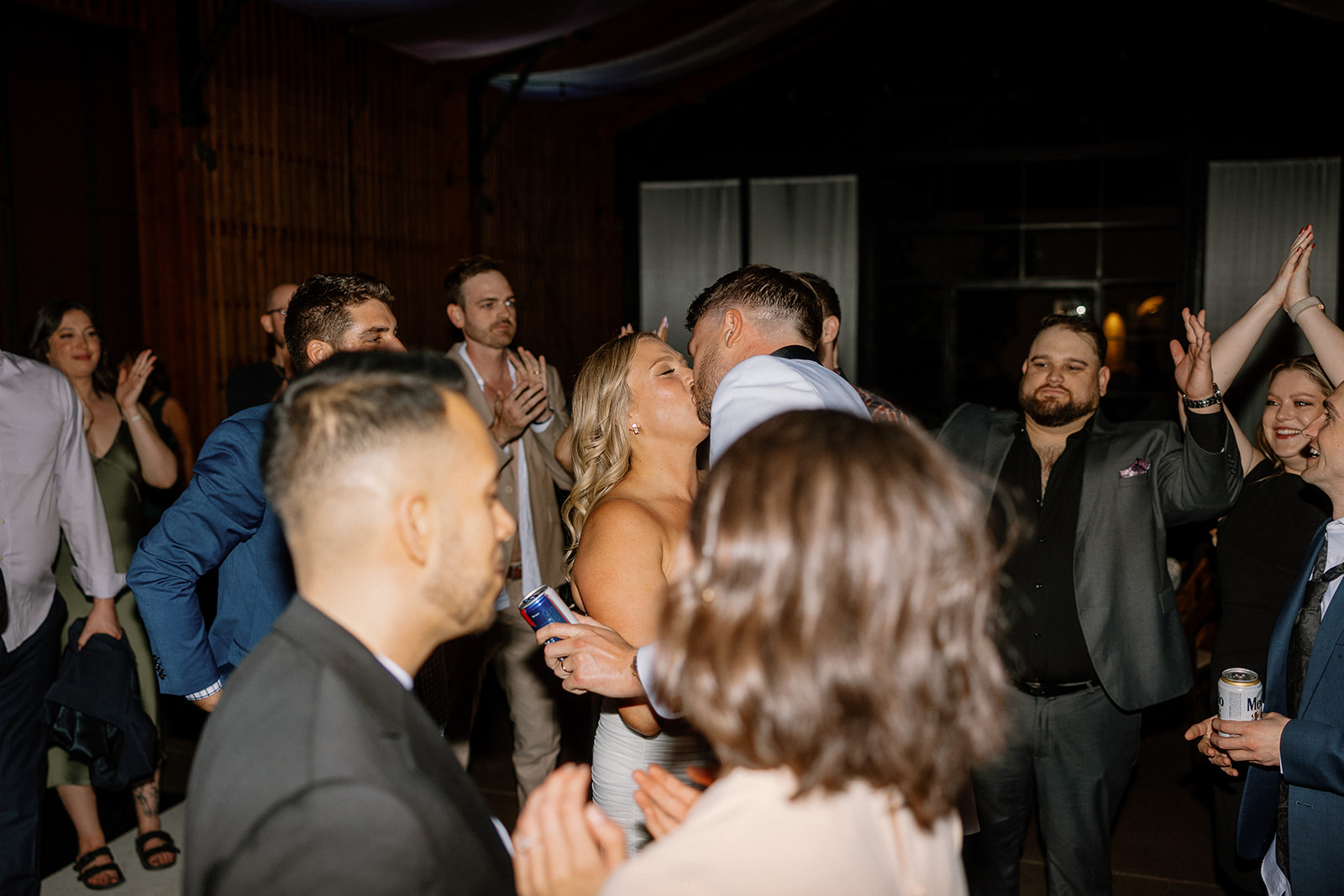 Groomsman lifted on another’s shoulders while dancing at a wedding reception with string lights and wooden interiors.
