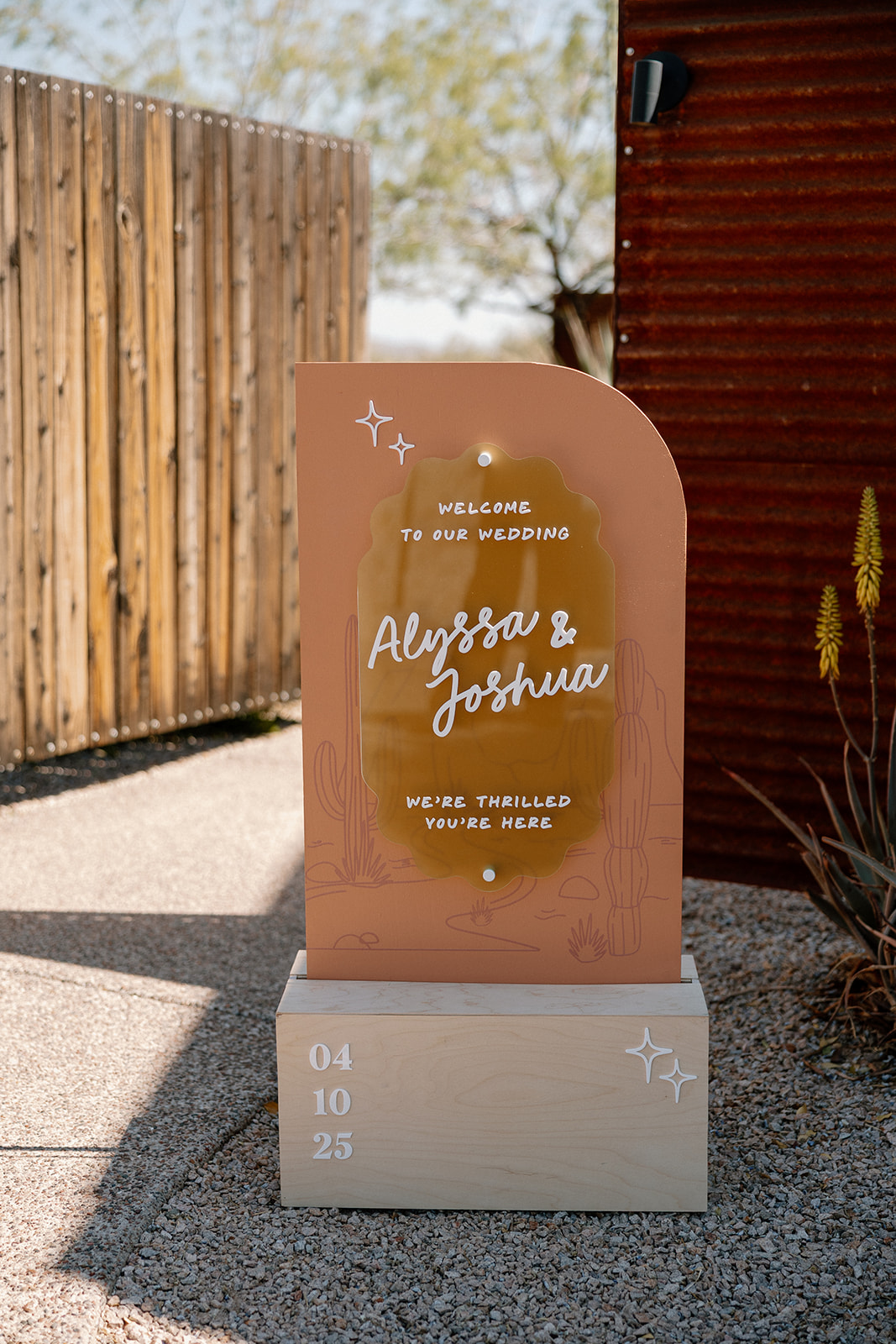 Terracotta and gold acrylic welcome sign with couple's names and wedding date, styled outside a modern desert venue.