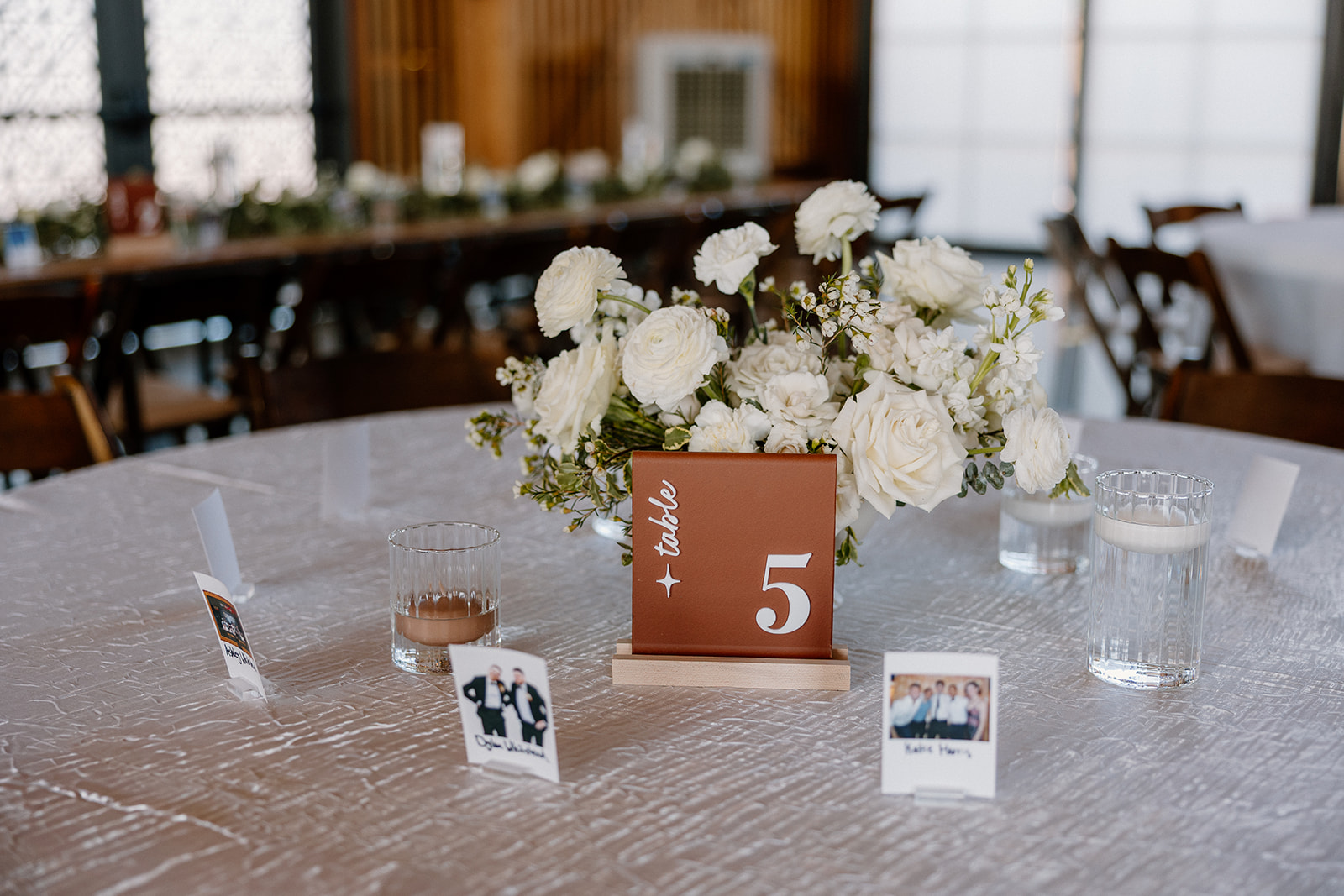 Terracotta table number centerpiece surrounded by white roses and guest photos at a reception table.