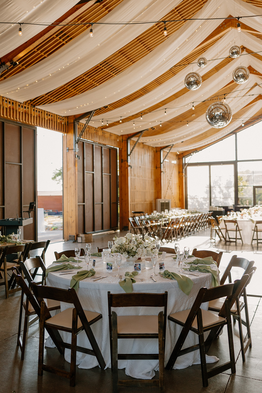 Round reception table set with white linens, green napkins, and candles under disco balls and sheer draping at a barn-style Arizona wedding venue.