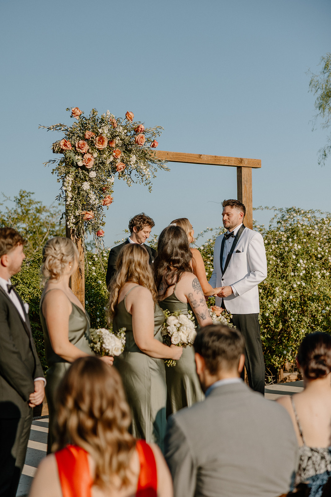Bride and groom exchanging vows under a wooden arch with bridesmaids in sage green dresses watching from the side.