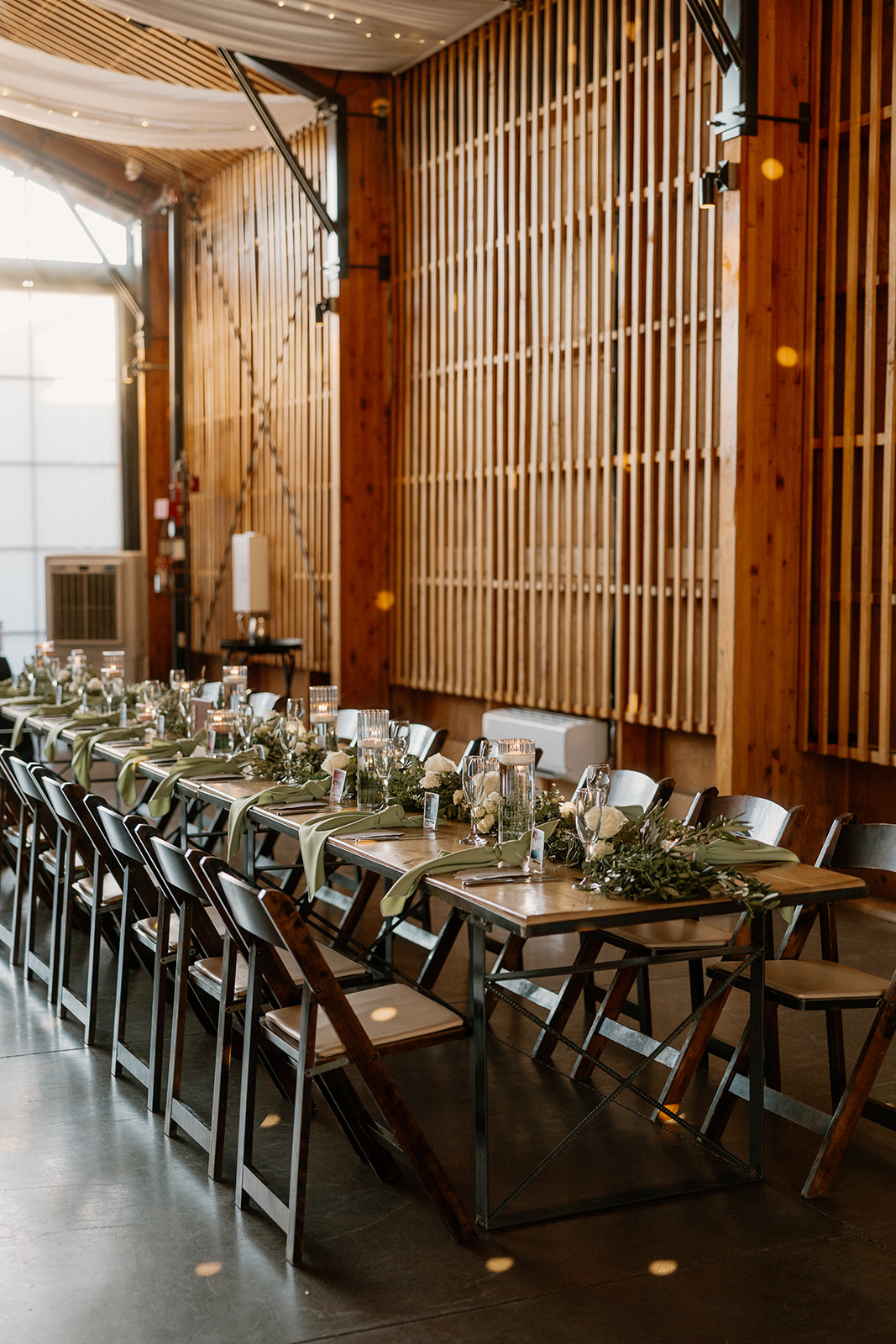 Long reception table with wooden chairs and greenery runners inside a modern barn venue.