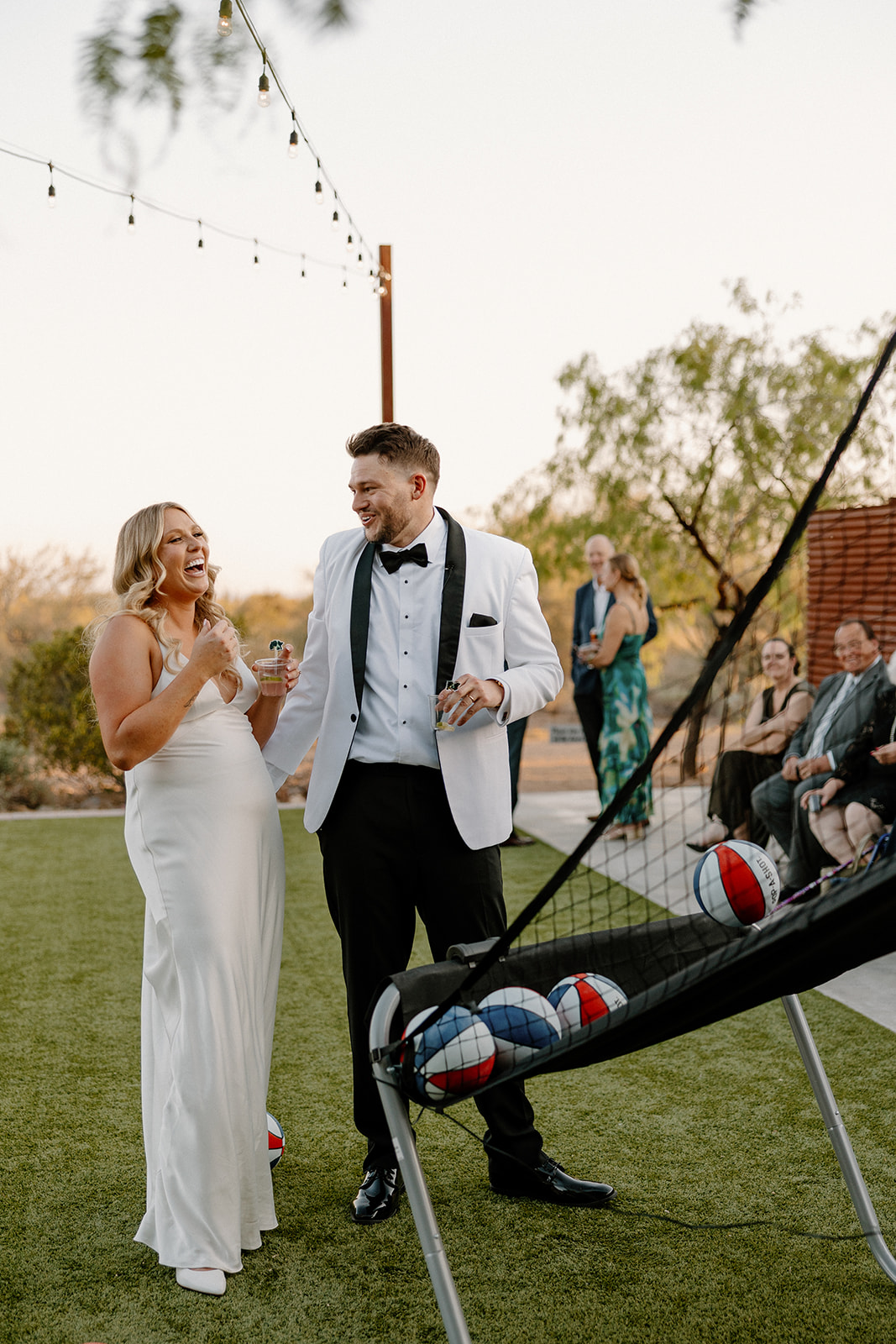 Bride and groom laughing together while playing a basketball arcade game during their wedding reception.
