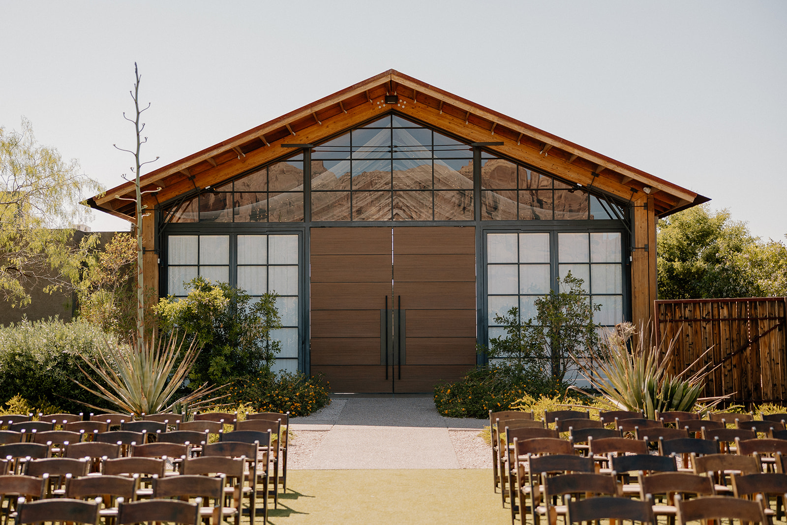 Outdoor ceremony setup with rows of wooden chairs facing a modern barn-style venue framed by desert greenery.
