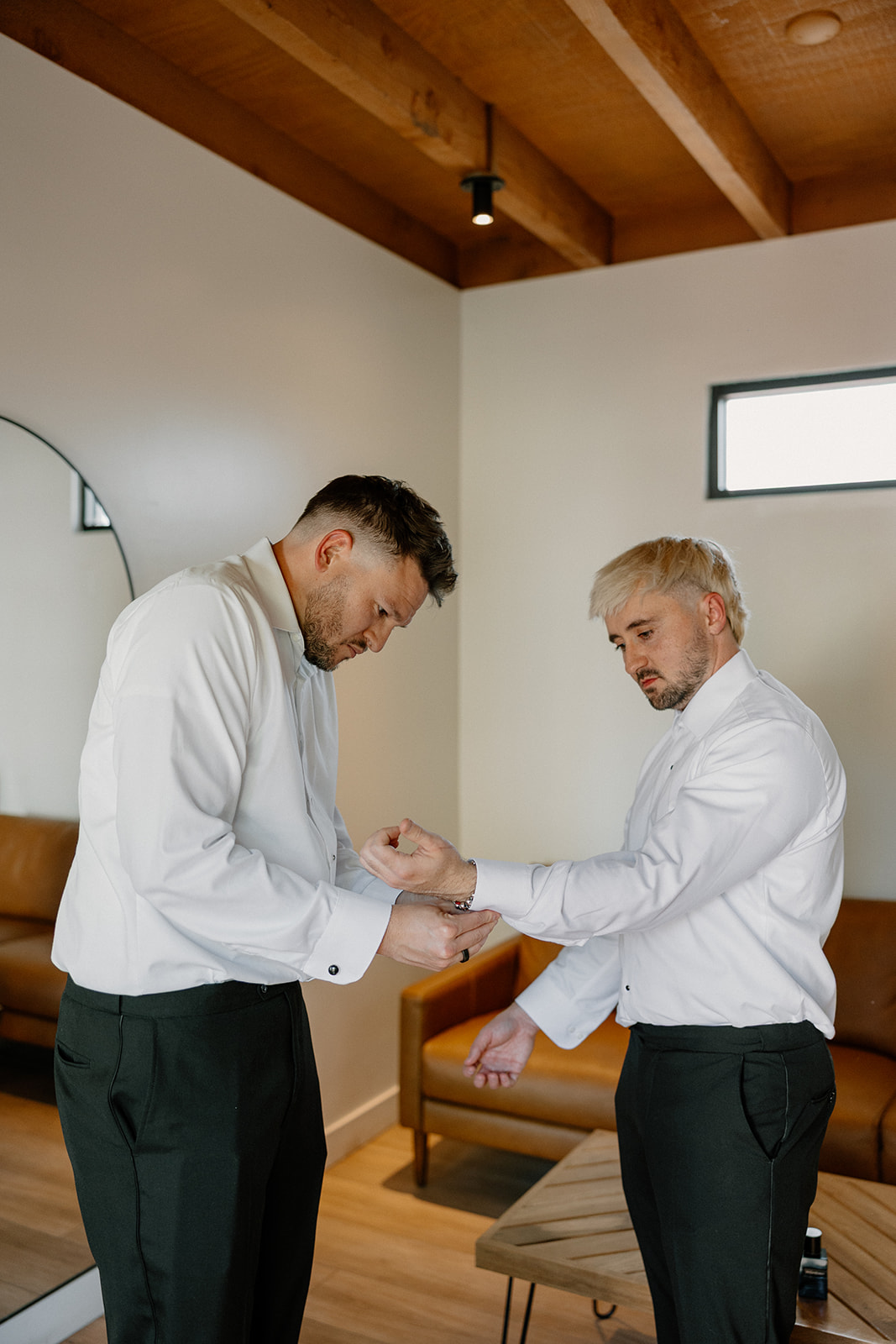Groom helps his best man with cufflinks in a modern prep space with natural light and wood accents.