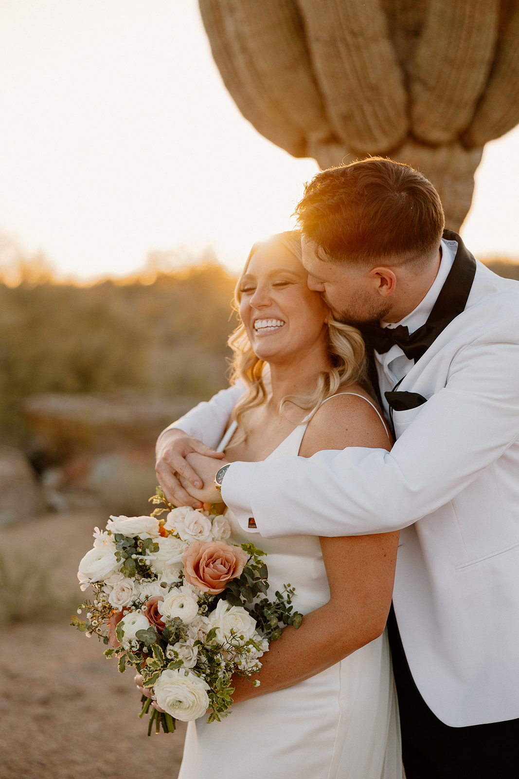 Groom kissing the bride on the cheek while she smiles with a sunset desert backdrop and a blooming bouquet.