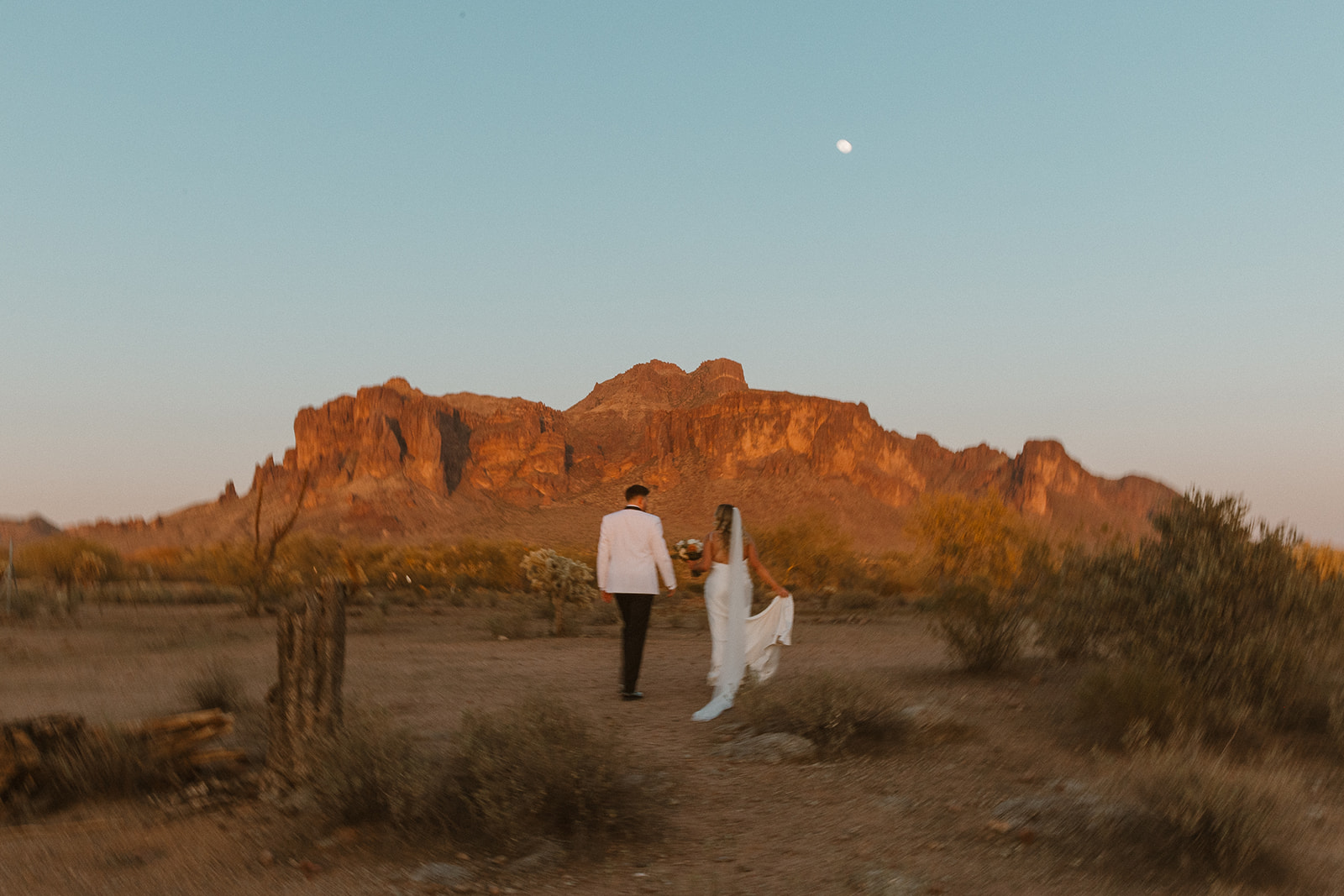 Bride and groom walking hand-in-hand through the desert at sunset, mountains glowing behind them under a soft moonlit sky.