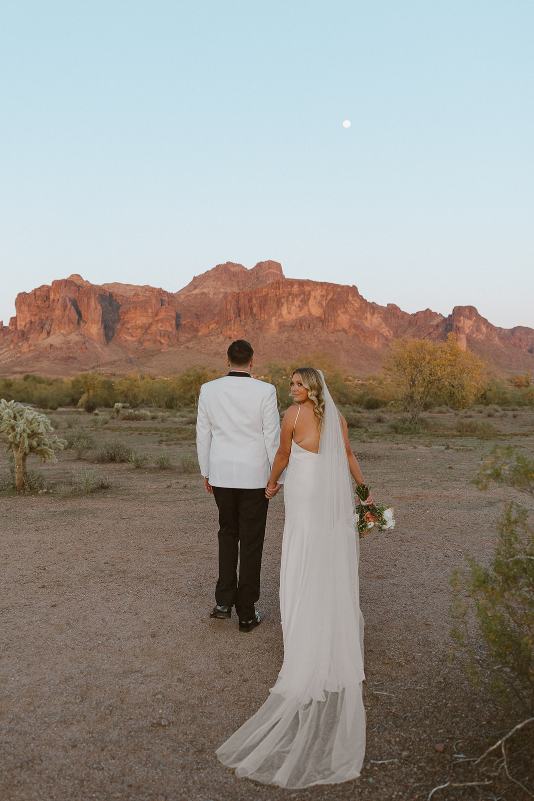 Bride and groom walking toward the Superstition Mountains at sunset, her veil trailing behind her at a wedding venue in Arizona.