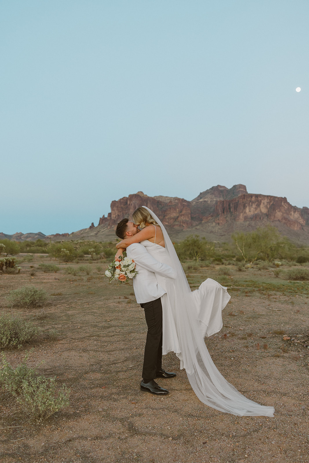 Groom lifts bride off the ground for a joyful kiss in the desert, surrounded by soft greenery and dramatic Arizona peaks.