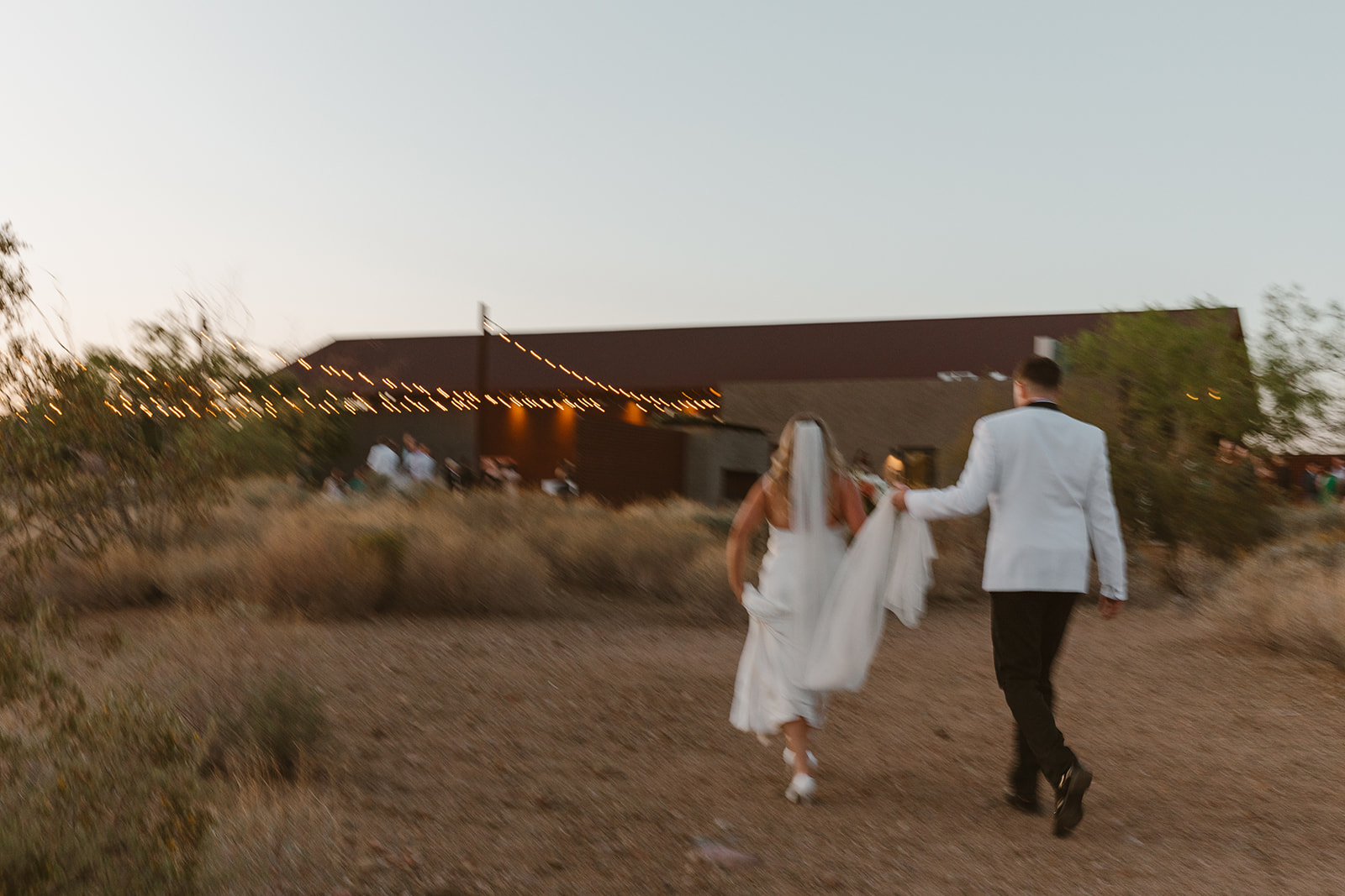 Newlyweds walking hand in hand through the desert path toward a warmly lit barn at sunset.