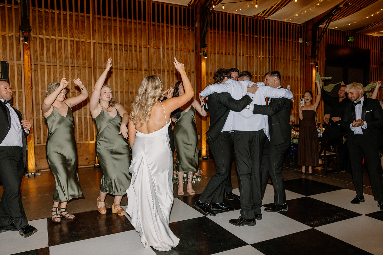 Bride and bridesmaids in sage dresses celebrating as groomsmen embrace in a group hug during a joyful wedding at one of Arizona’s standout venues.