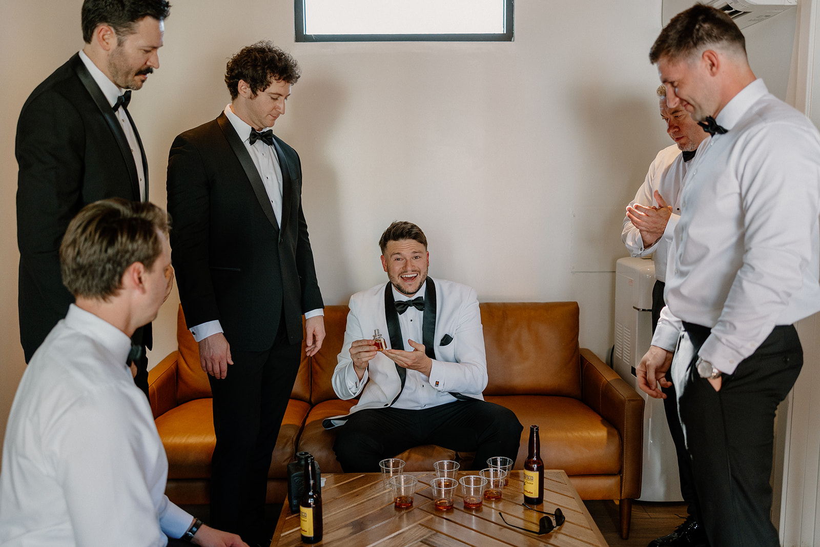 Groom sits on a leather couch, smiling and holding a glass while surrounded by groomsmen in a private lounge before the ceremony.
