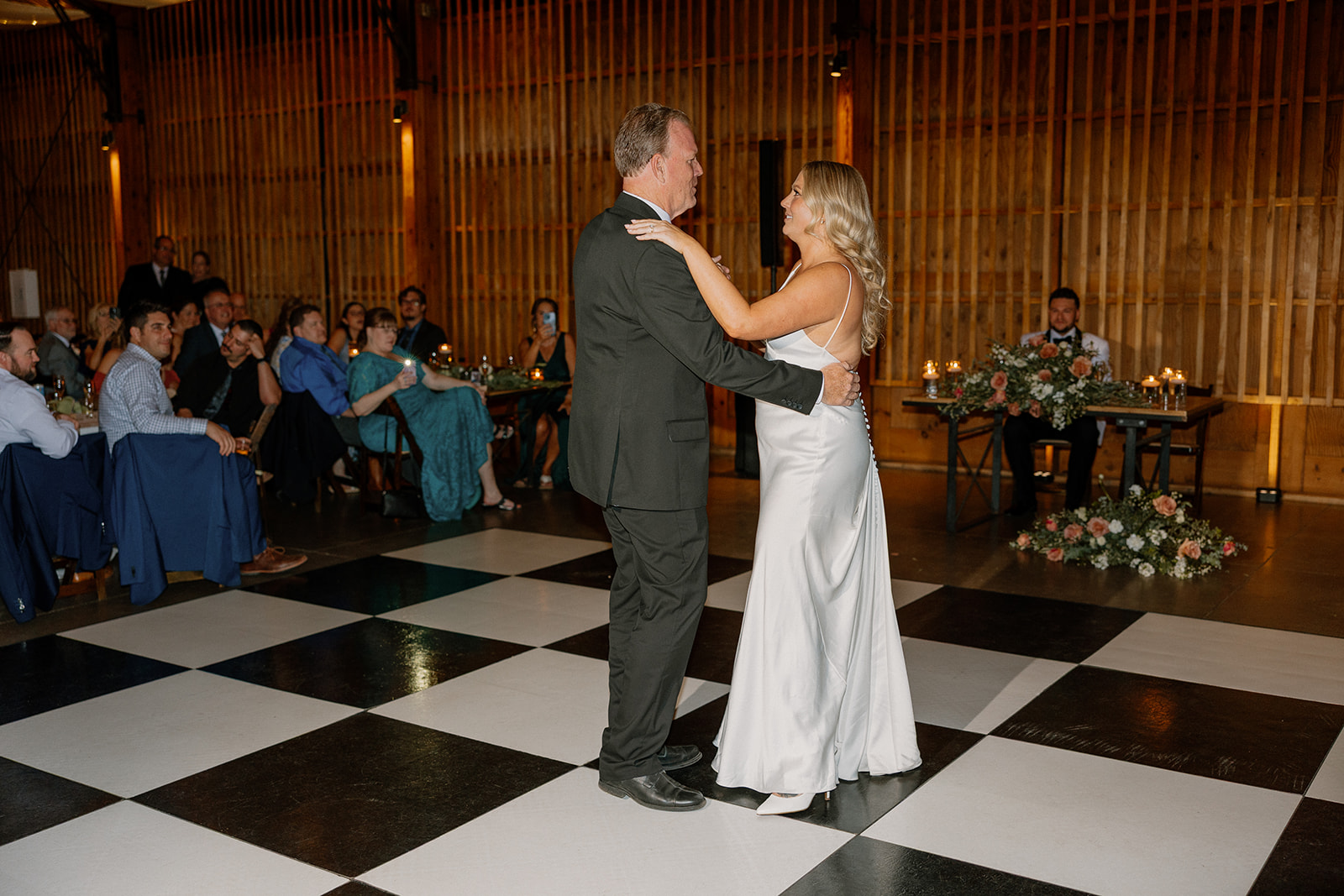 Father-daughter dance on a checkered floor, with guests watching from their seats in a warmly lit venue.
