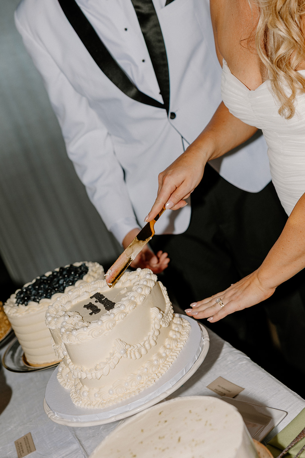 Bride cutting into a white wedding cake decorated with piped frosting and a custom topper, while the groom watches.