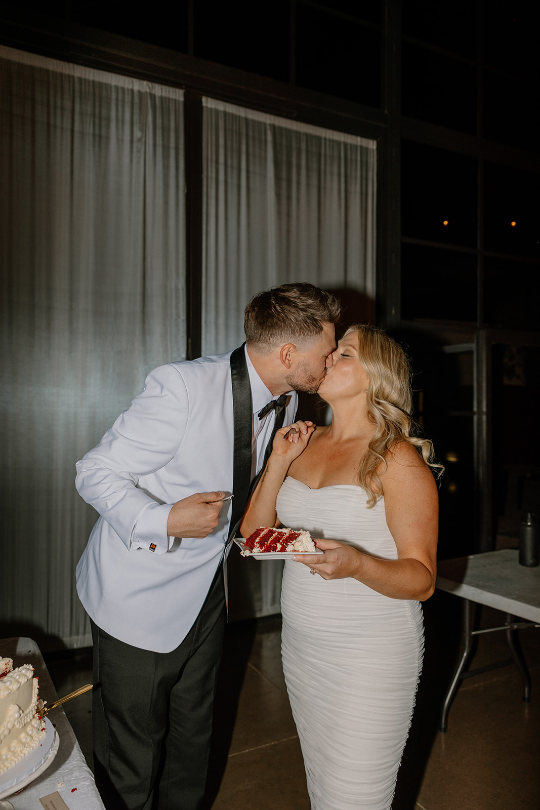 Groom kissing the bride as she holds a plate of cake, sharing a sweet moment during their reception.