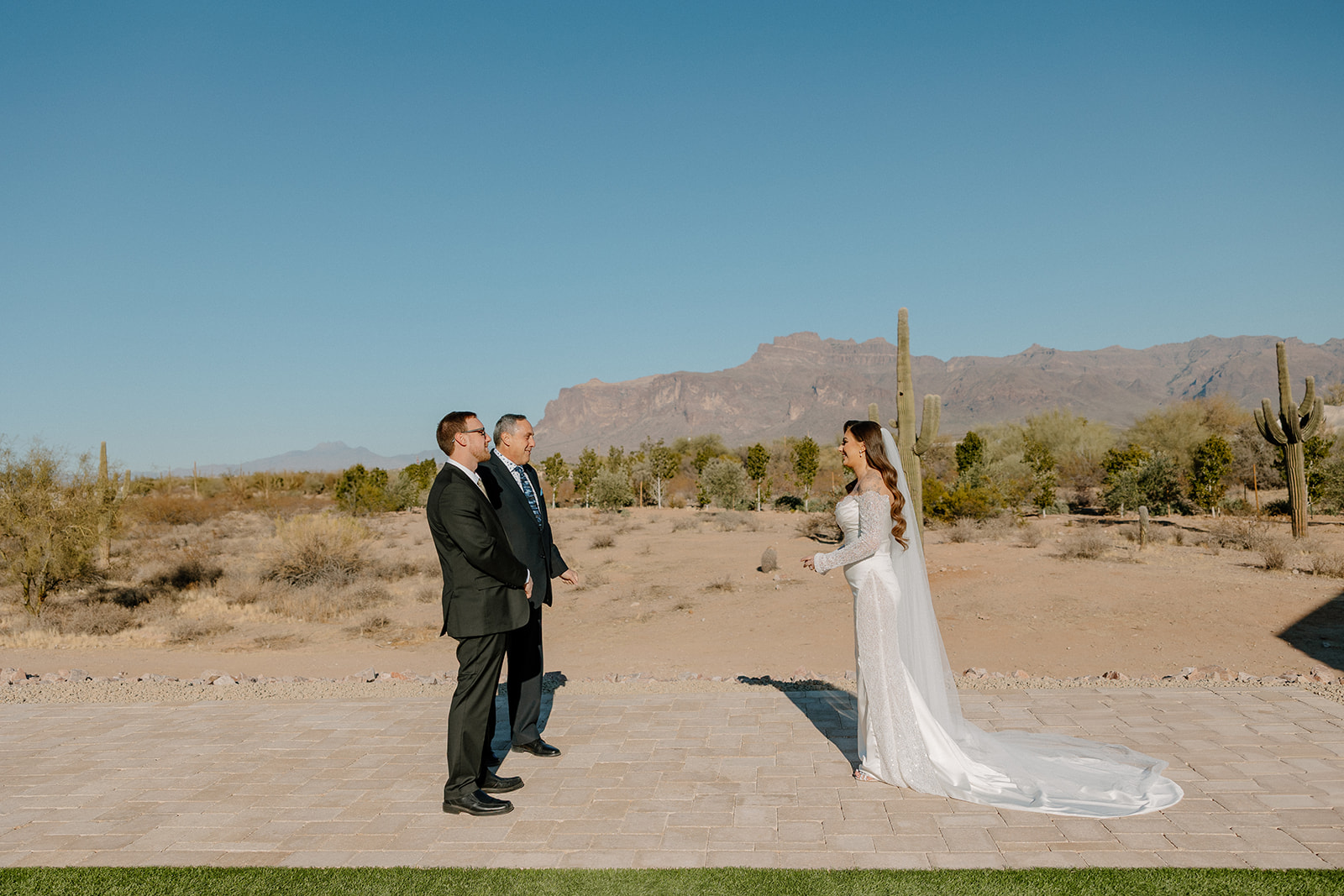 Bride facing two officiants during a wedding ceremony in the open air, with cacti and mountains in the background at a desert view wedding venue.
